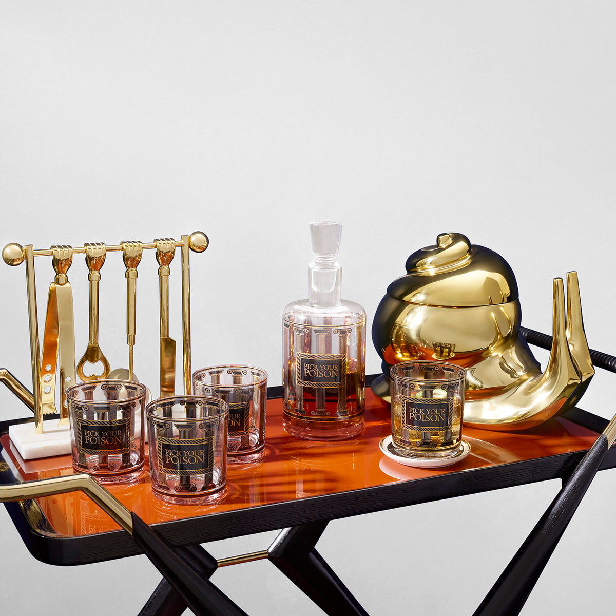 A luxury bar cart styled with a gold teapot, a gold decanter labeled RASON, two gold and clear tumblers, a gold and clear glassware holder, and a copper-toned wooden tray on a black-framed cart.