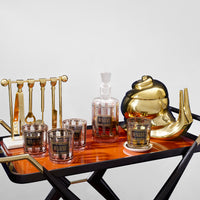 A luxury bar cart styled with a gold teapot, a gold decanter labeled RASON, two gold and clear tumblers, a gold and clear glassware holder, and a copper-toned wooden tray on a black-framed cart.