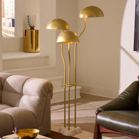 A modern three-arm floor lamp with a brushed gold finish and dome-shaped shades, styled in a contemporary living room next to a textured beige sofa and a dark leather chair.
