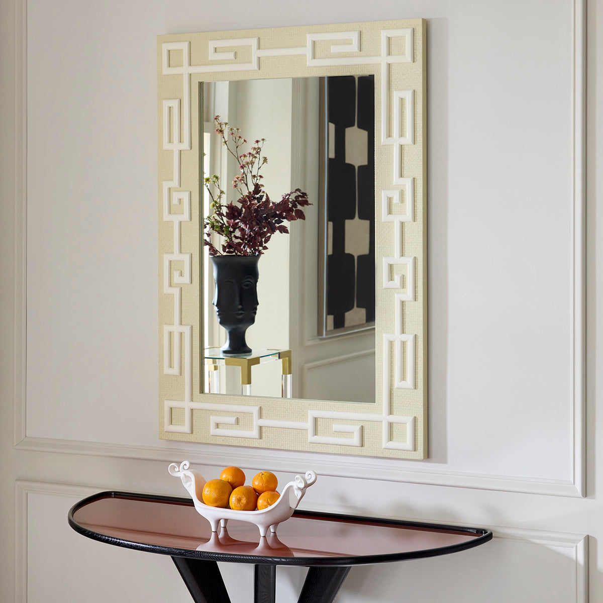 A rectangular wall mirror with an ornate cream and white Greek key geometric frame, mounted on a white paneled wall above a console table with a bowl of oranges.