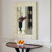 A rectangular wall mirror with an ornate cream and white Greek key geometric frame, mounted on a white paneled wall above a console table with a bowl of oranges.
