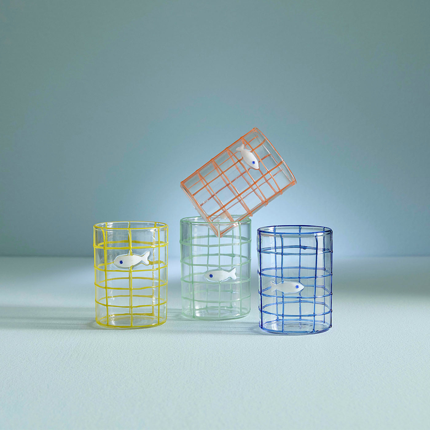 Three glass tumblers with wireframe designs and small fish figures on a teal background
