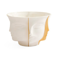 A sculptural ceramic bowl featuring a split-face design with one half showing a profile of a face in profile and the other half showing a profile of a face facing forward, in glossy off-white with a warm gold interior accent.