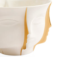 A close-up of a sculptural ceramic planter or bowl with a white face design and metallic gold accents, shown against a plain white background.