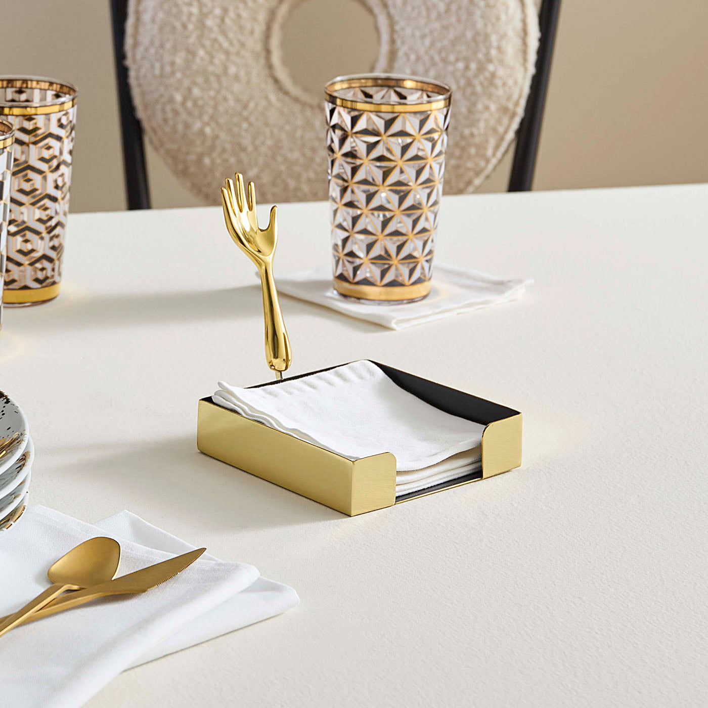 Dining table setting with gold cutlery, geometric-patterned glasses, and a napkin holder.