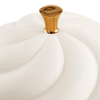 A close-up of a white ceramic container lid with a sculpted, swirled design and a polished gold knob on a white background.