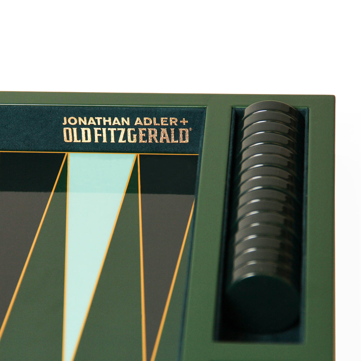 Close-up of a Jonathan Adler + Old Fitzgerald board game box corner, showing dark green packaging with gold lettering and a stack of round black pieces in a side slot.