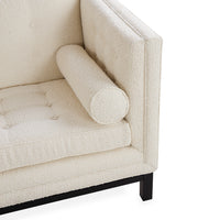 Close-up of a cream bouclé armchair corner with a cylindrical bolster armrest, tufted seat cushion, and black wooden legs on a white background.