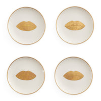 Four round white plates arranged in a 2x2 grid on a white background, each with a thin gold rim and a gold lips motif centered on the plate.