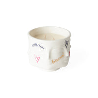 A white ceramic candle in a sculpted skull-shaped container, decorated with colorful hand-drawn hearts, eyelashes, and the word 'biscuit' in gold script, set against a plain white background.