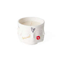 A white ceramic candle holder sculpted in the shape of a face, containing a white wax candle with a single wick. The ceramic features hand-painted doodles in black, gold, and red, including the word 'biscus!' in gold script, a black heart outline, and red lips.
