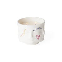 A white ceramic candle holder shaped like a stylized face, with two lit candles inside. The face features minimalist hand-drawn style illustrations in gold, navy blue, and pink, including the words 'Be Kind' in gold script.