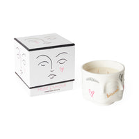 A white ceramic candle jar with a sculpted face design and pink heart details, shown next to its white cube-shaped packaging box featuring minimalist face line art.