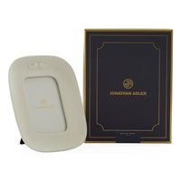 A cream-colored Joseph Louis Mount picture frame with a small gold Jonathan Adler logo, shown next to its dark navy and gold presentation box on a white background.