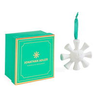 Jonathan Adler porcelain ornament shaped like a snowflake with a green ribbon loop, shown next to its signature green and gold gift box on a white background.