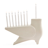 A white ceramic menorah shaped like a bird, holding nine white candles with lit flames, photographed on a seamless white background.