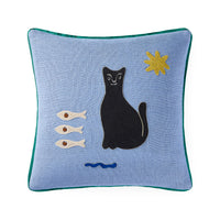 A square light blue throw pillow with a green piped edge, featuring an embroidered design of a black cat, three white fish, a blue wave, and a yellow star on a white background.