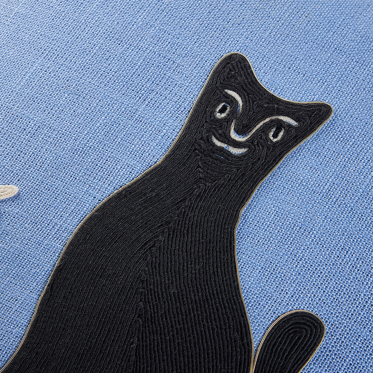 A close-up of a black embroidered cat patch with white facial details, placed on a textured light blue fabric background.