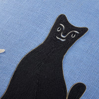 A close-up of a black embroidered cat patch with white facial details, placed on a textured light blue fabric background.