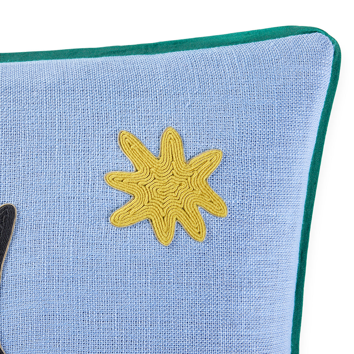 A close-up of a light blue textured fabric pillow with a dark green piped edge and a yellow embroidered starburst motif on the front.