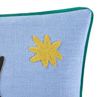 A close-up of a light blue textured fabric pillow with a dark green piped edge and a yellow embroidered starburst motif on the front.