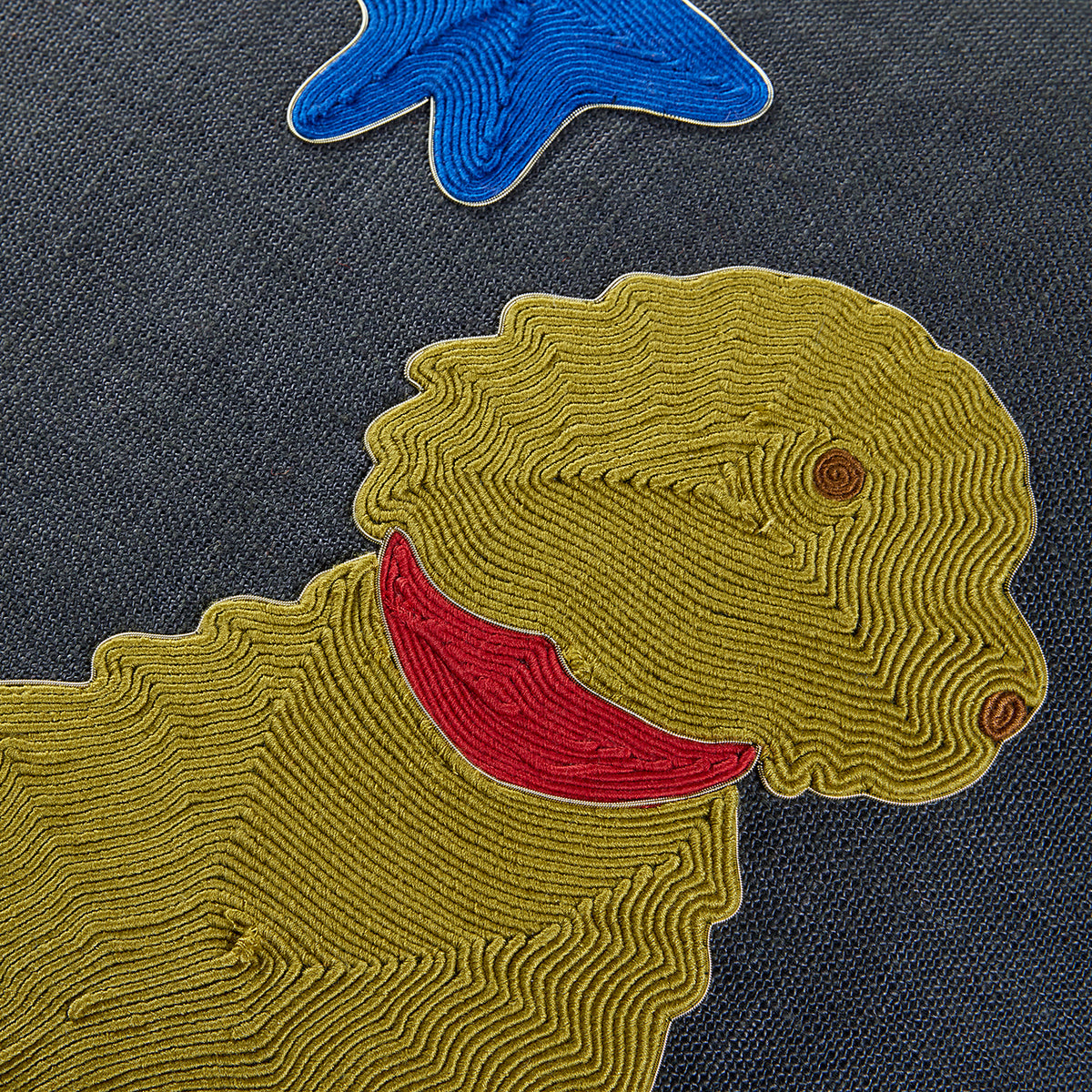 Close-up of a textured embroidered patch featuring a yellow dog with a red collar on a dark gray woven fabric background, with part of a blue star patch visible at the top.