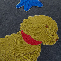 Close-up of a textured embroidered patch featuring a yellow dog with a red collar on a dark gray woven fabric background, with part of a blue star patch visible at the top.