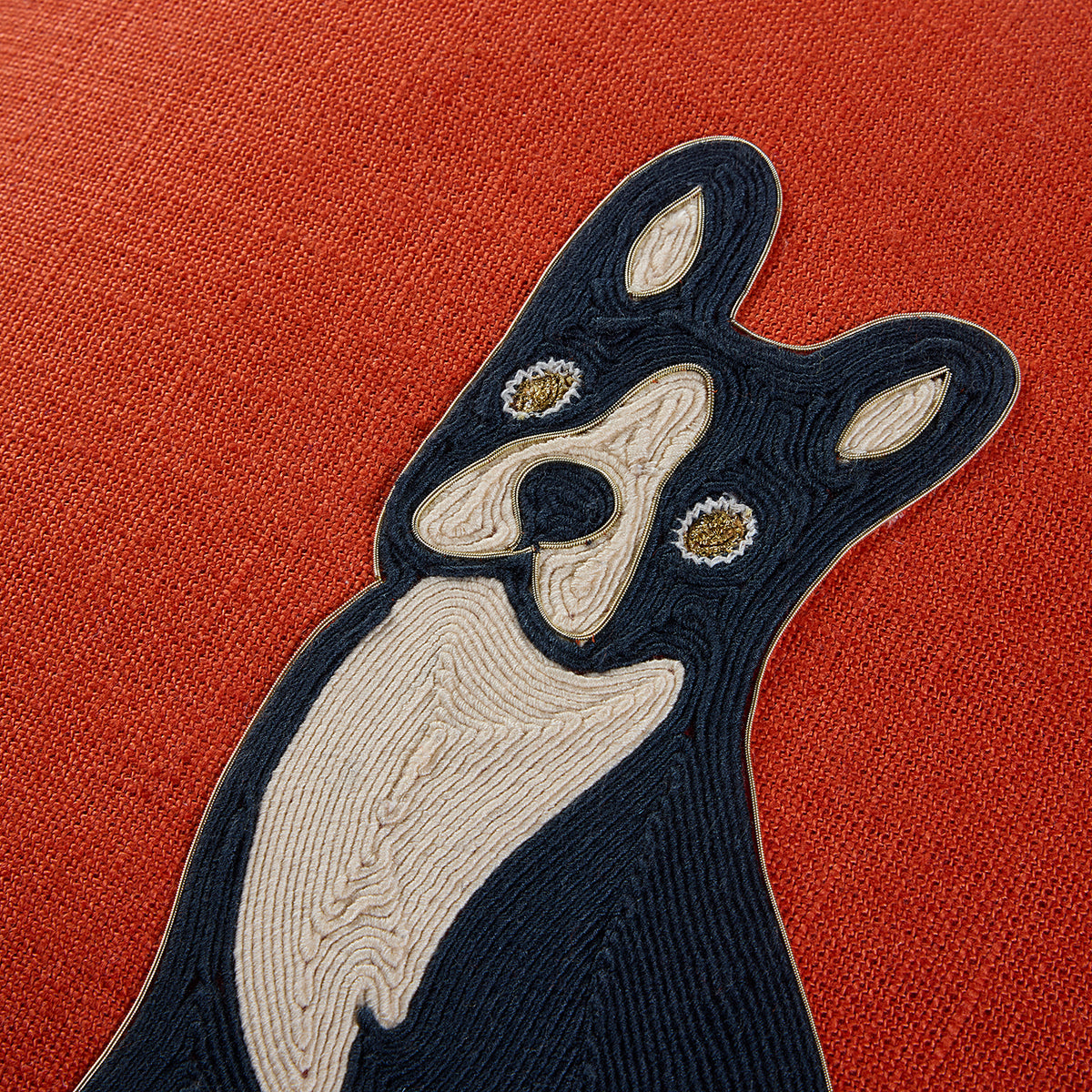 A close-up of a textured red-orange fabric featuring a stylized embroidered dog motif in dark navy and cream with small gold and white eye details.