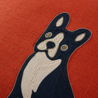 A close-up of a textured red-orange fabric featuring a stylized embroidered dog motif in dark navy and cream with small gold and white eye details.