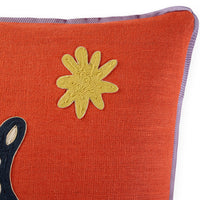 Close-up of a textured orange throw pillow with a yellow embroidered starburst motif and a purple border, on a white background.