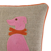 Close-up of a textured beige throw pillow with a pink embroidered dog and an orange collar, finished with an orange piped edge.