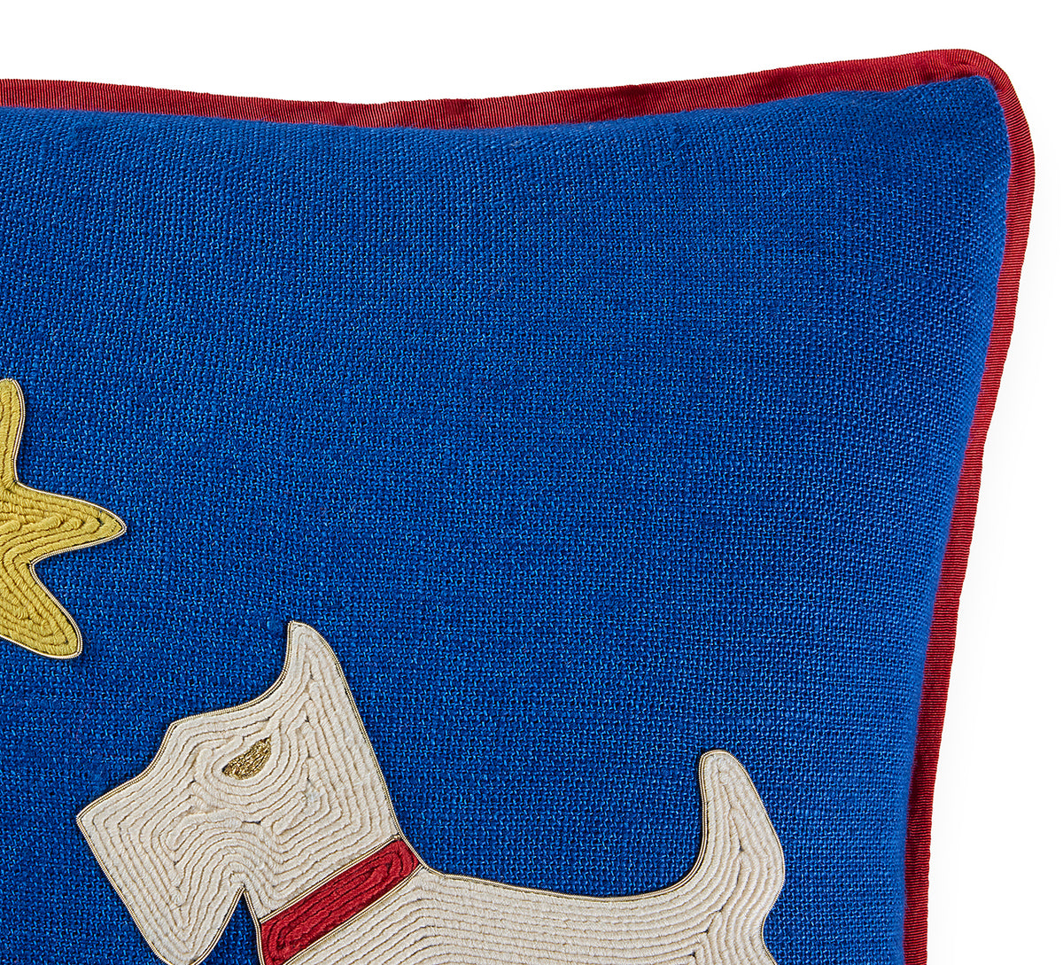 Close-up of a blue embroidered throw pillow featuring a white dog with a red collar and a yellow star, finished with a red velvet trim.