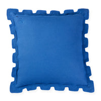 A square royal blue throw pillow with a decorative scalloped or wavy piped border, shown against a plain white background. A small circular monogram logo is embroidered in the top-left corner.