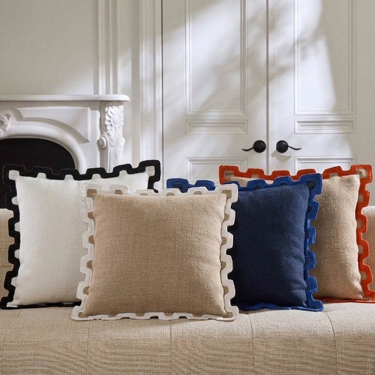 A set of square decorative throw pillows with scalloped edges arranged on a beige upholstered sofa in a bright room. The visible pillows include white, beige, navy blue, and rust orange in different color combinations.