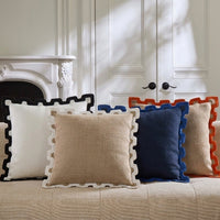 A set of square decorative throw pillows with scalloped edges arranged on a beige upholstered sofa in a bright room. The visible pillows include white, beige, navy blue, and rust orange in different color combinations.