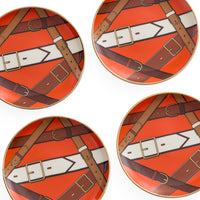 Four round decorative plates arranged in a 2x2 grid, featuring a repeating pattern of brown leather straps and buckles over a bright orange background with white arrow-like shapes.
