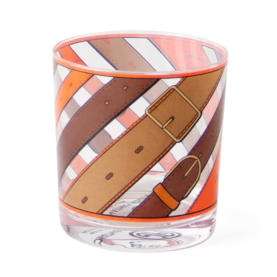 A clear glass tumbler featuring a printed design of overlapping leather straps in brown, tan, and orange with white and orange diagonal stripes, shown on a white background.