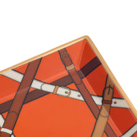 Close-up of a square decorative tray with rounded corners, featuring an orange background and an illustration of brown and white leather straps with buckles, finished with a gold-toned rim.