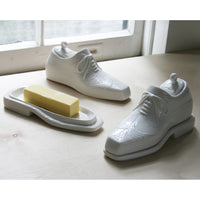 White ceramic shoes and a bar of soap on a light wooden surface with a window in the background.