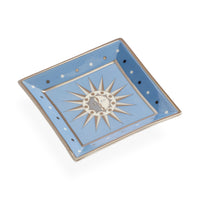 A square ceramic dish with a light blue glaze, featuring a central silver sunburst motif with a small moon and stars, and silver-rimmed edges with white and dark blue polka dots.