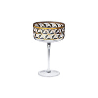 A clear stemmed coupe glass with a geometric faceted pattern in black, white, and gold on the bowl, set against a plain white background.