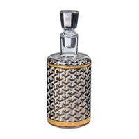 A clear glass decanter with a faceted stopper, featuring a cylindrical body decorated with a black, white, and gold geometric pattern and gold bands near the top and bottom, shown on a plain white background.