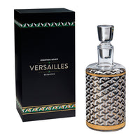 Jonathan Adler Versailles Decanter by Jonathan Adler, featuring a clear glass decanter with a black, white, and gold geometric pattern, shown next to its black presentation box with green and gold accents.
